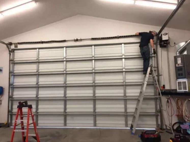 A man is repairing a garage door inside a garage, focused on his task with tools nearby.