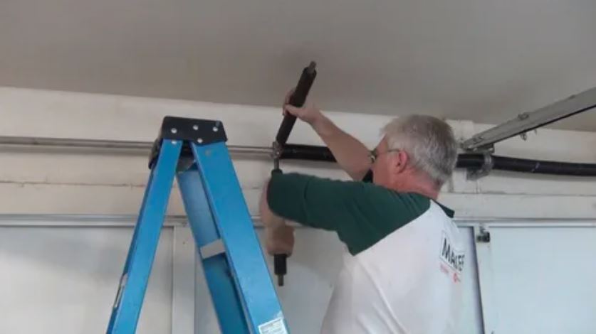 A man repairs a garage door while using a ladder for support and access to the upper mechanism.